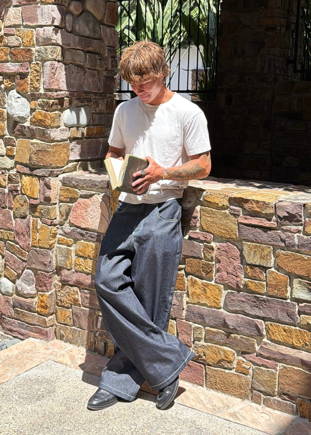A man wearing Divine 144 baggy raw dark indigo denim jeans with a wide straight-leg fit, leaning against a stone wall.