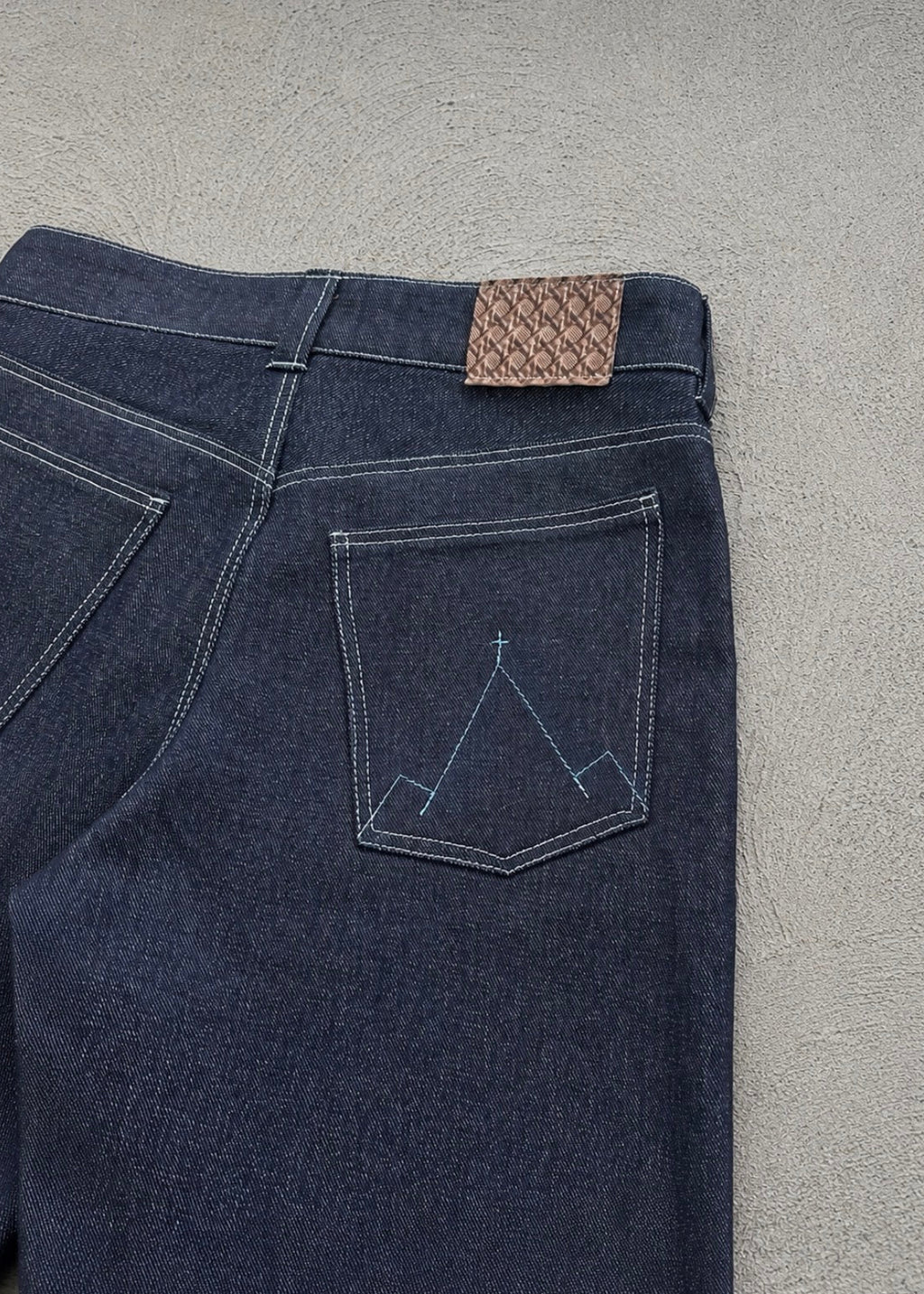 Divine 144 dark indigo raw denim jeans featuring a leather patch and signature mountain embroidery on the back pocket.